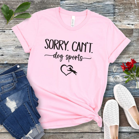 A pink T-shirt with the text 'SORRY, CAN'T dog sports' and a heart graphic with dog leaping through it under the text, displayed on a wooden background with a pair of jeans and white shoes.