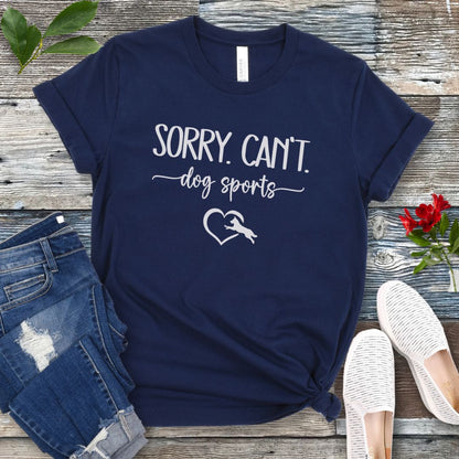A navy T-shirt with the text 'SORRY, CAN'T dog sports' and a heart graphic with dog leaping through it under the text, displayed on a wooden background with a pair of jeans and white shoes.
