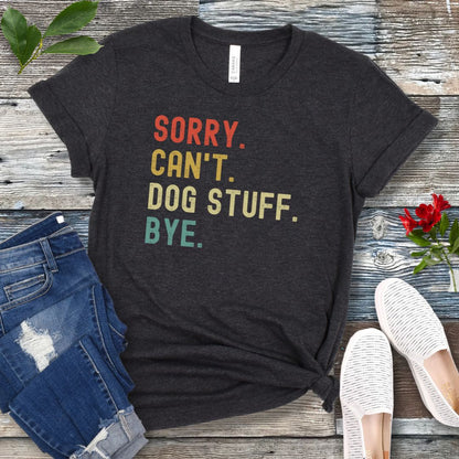 A dark grey unisex short sleeve T-shirt with colorful stacked text 'SORRY. CAN'T. DOG STUFF. BYE.', presented on a wooden surface with jeans and shoes.