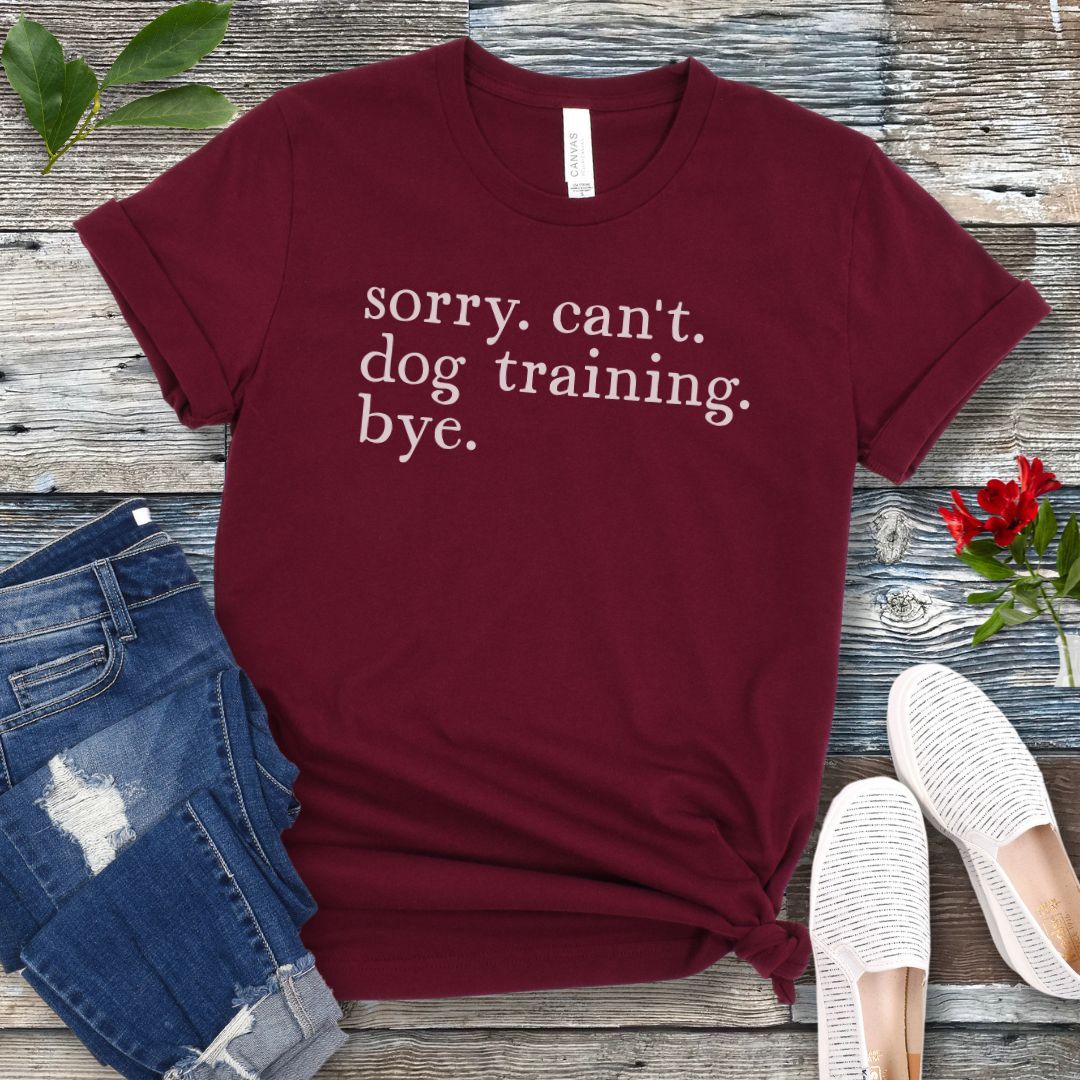 Maroon t-shirt with 'sorry. can't. dog training. bye.' text on a wooden surface with jeans and shoes.