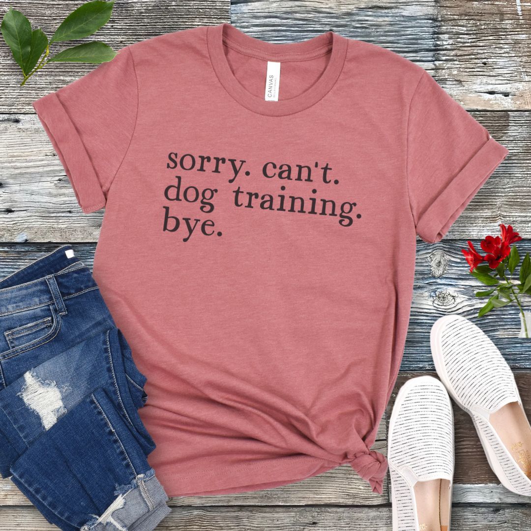 Mauve t-shirt with 'sorry. can't. dog training. bye.' text on a wooden surface with jeans and shoes.
