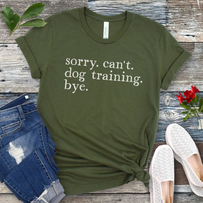 Green t-shirt with 'sorry. can't. dog training. bye.' text on a wooden surface with jeans and shoes.