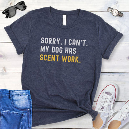 Heather navy t-shirt with 'SORRY, CAN'T. MY DOG HAS SCENT WORK' text on a wooden surface with jeans and shoes.