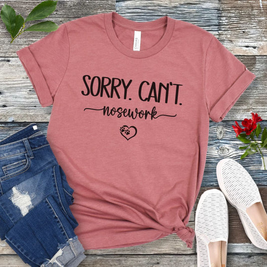 Mauve dog sports t-shirt with 'Sorry. Can't. Nosework' text on a wooden surface with jeans and shoes.