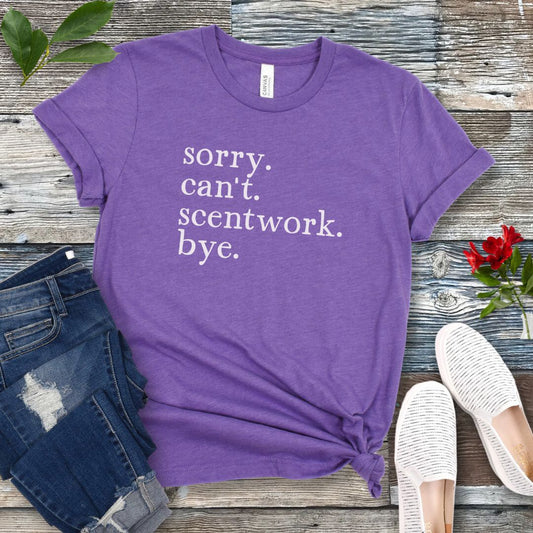 Purple dog sports t-shirt with text 'sorry. can't. scentwork. bye.' on a wooden surface with jeans and shoes.