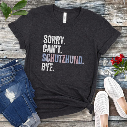 Dark grey t-shirt with 'SORRY. CAN'T. SCHUTZHUND. BYE' in colorful text, presented on a wooden surface with jeans and shoes.