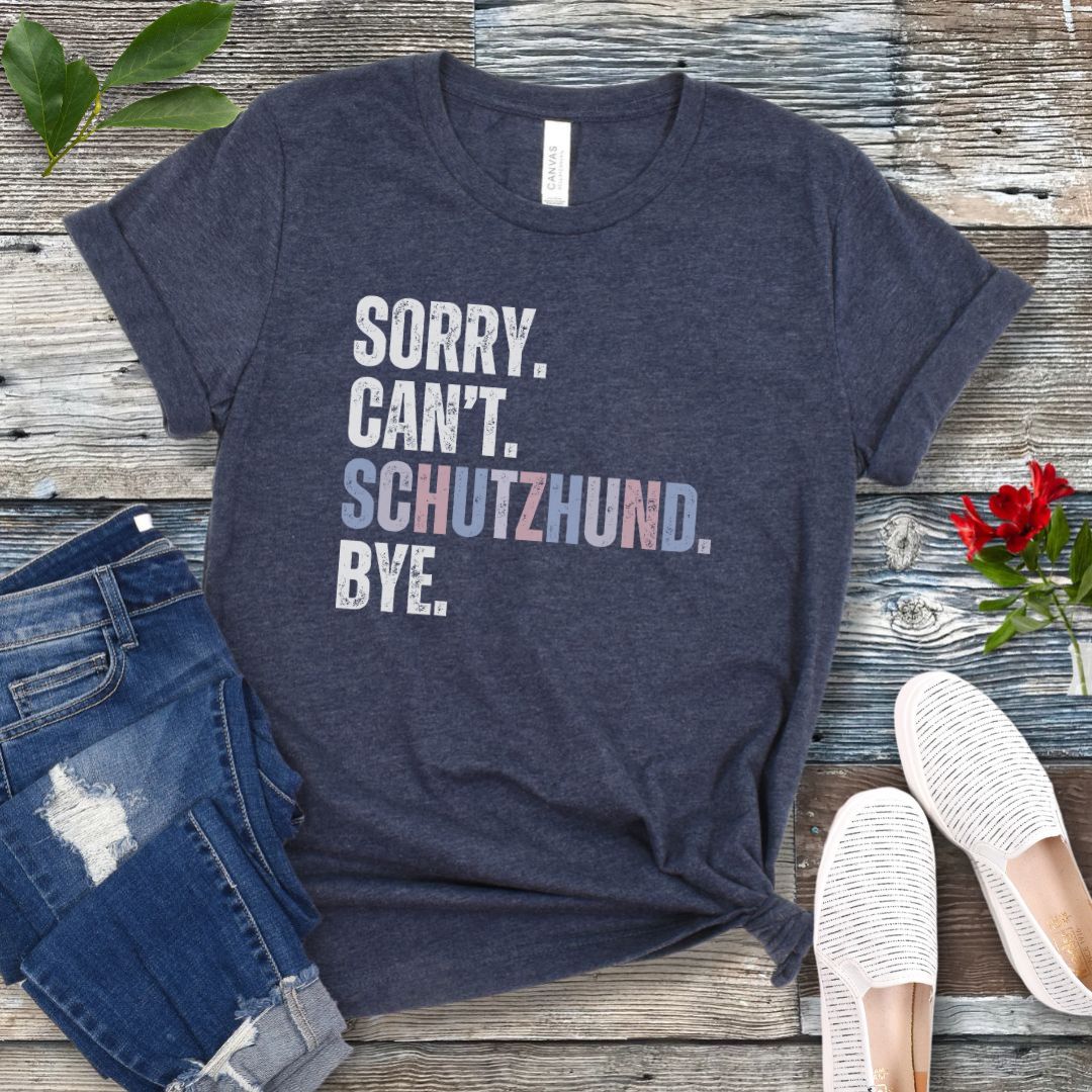 Heather navy blue t-shirt with 'SORRY. CAN'T. SCHUTZHUND. BYE' in colorful text, presented on a wooden surface with jeans and shoes.