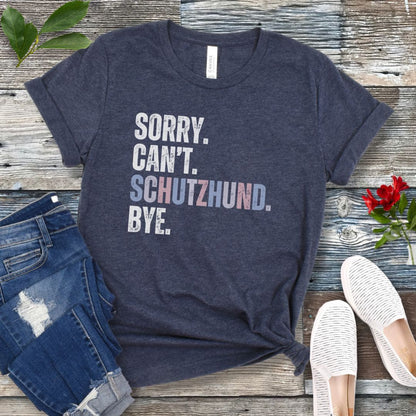 Heather navy blue t-shirt with 'SORRY. CAN'T. SCHUTZHUND. BYE' in colorful text, presented on a wooden surface with jeans and shoes.