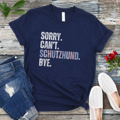 Navy blue t-shirt with 'SORRY. CAN'T. SCHUTZHUND. BYE' in colorful text, presented on a wooden surface with jeans and shoes.