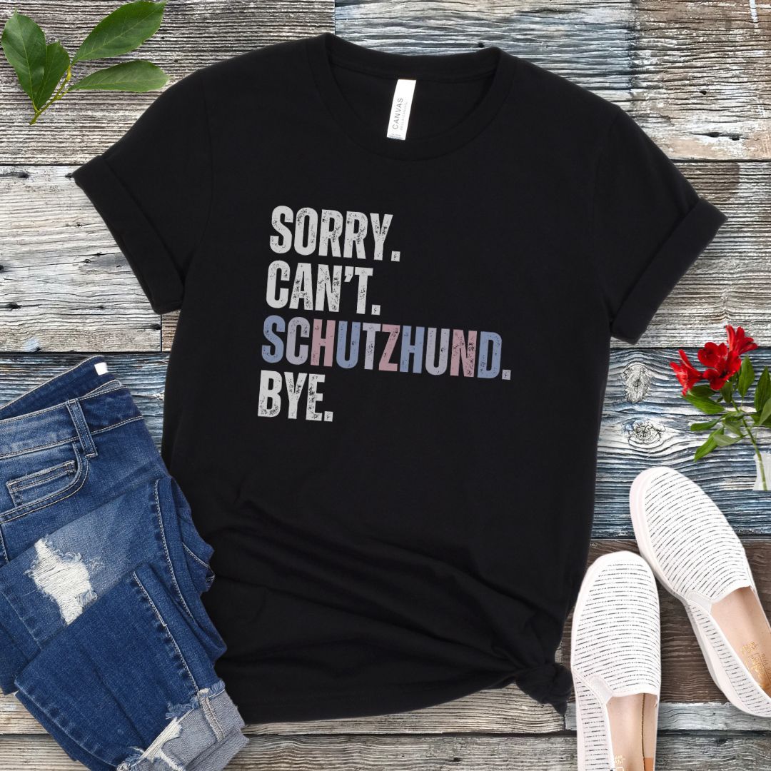 Black t-shirt with 'SORRY. CAN'T. SCHUTZHUND. BYE' in colorful text, presented on a wooden surface with jeans and shoes.