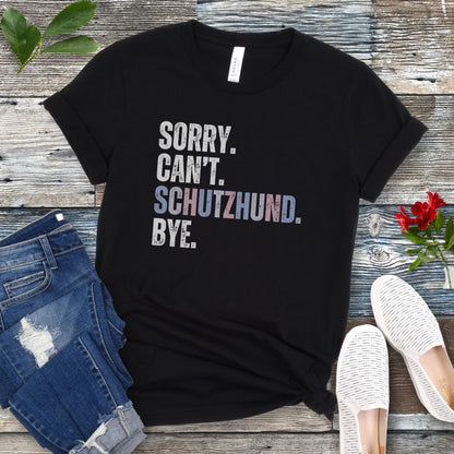 Black t-shirt with 'SORRY. CAN'T. SCHUTZHUND. BYE' in colorful text, presented on a wooden surface with jeans and shoes.