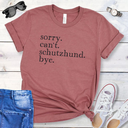 Mauve t-shirt with 'sorry, can't, Schutzhund, bye' text on a wooden surface with jeans and shoes.