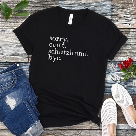 Black t-shirt with 'sorry, can't, Schutzhund, bye' text on a wooden surface with jeans and shoes.