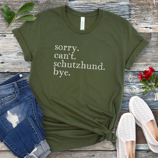 Military green t-shirt with text 'sorry. can't. schutzhund. bye.' on a wooden surface with jeans and shoes.