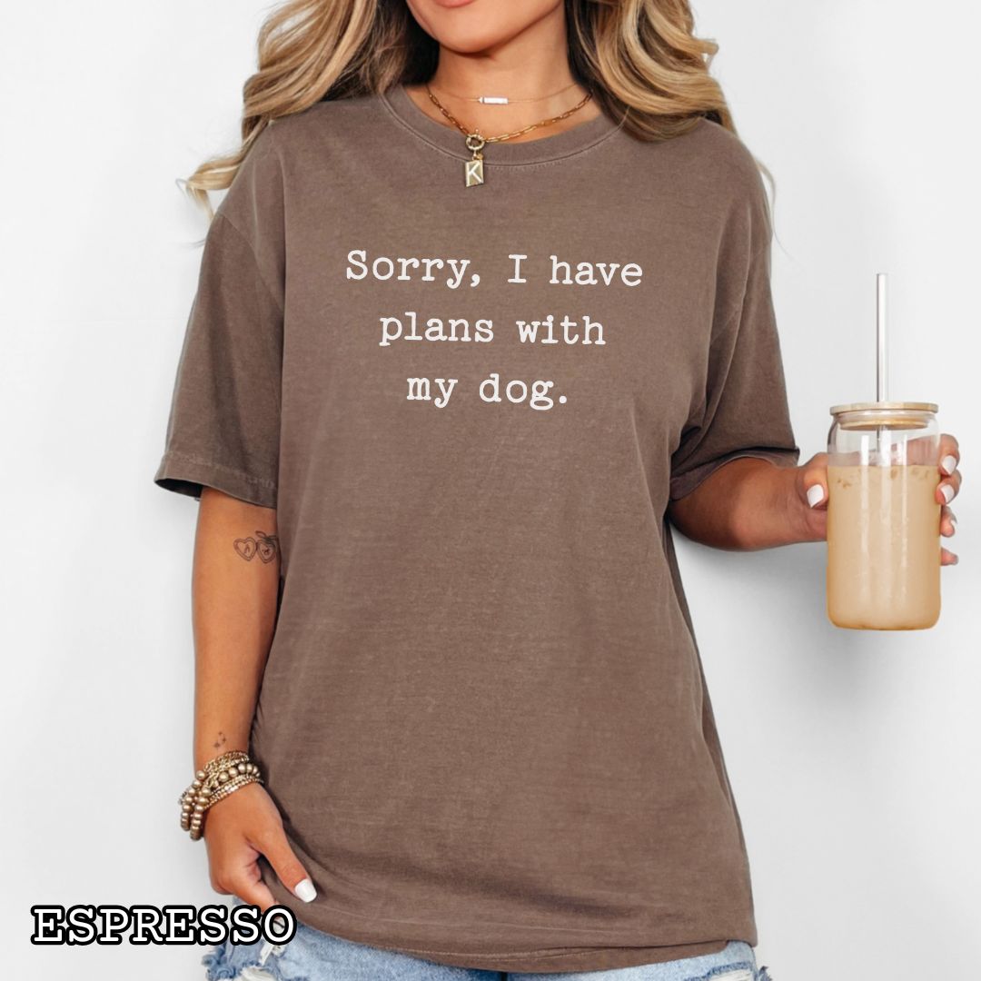 Person wearing a brown Comfort Colors dog lover t-shirt with 'Sorry, I have plans with my dog.' text, holding a drink.