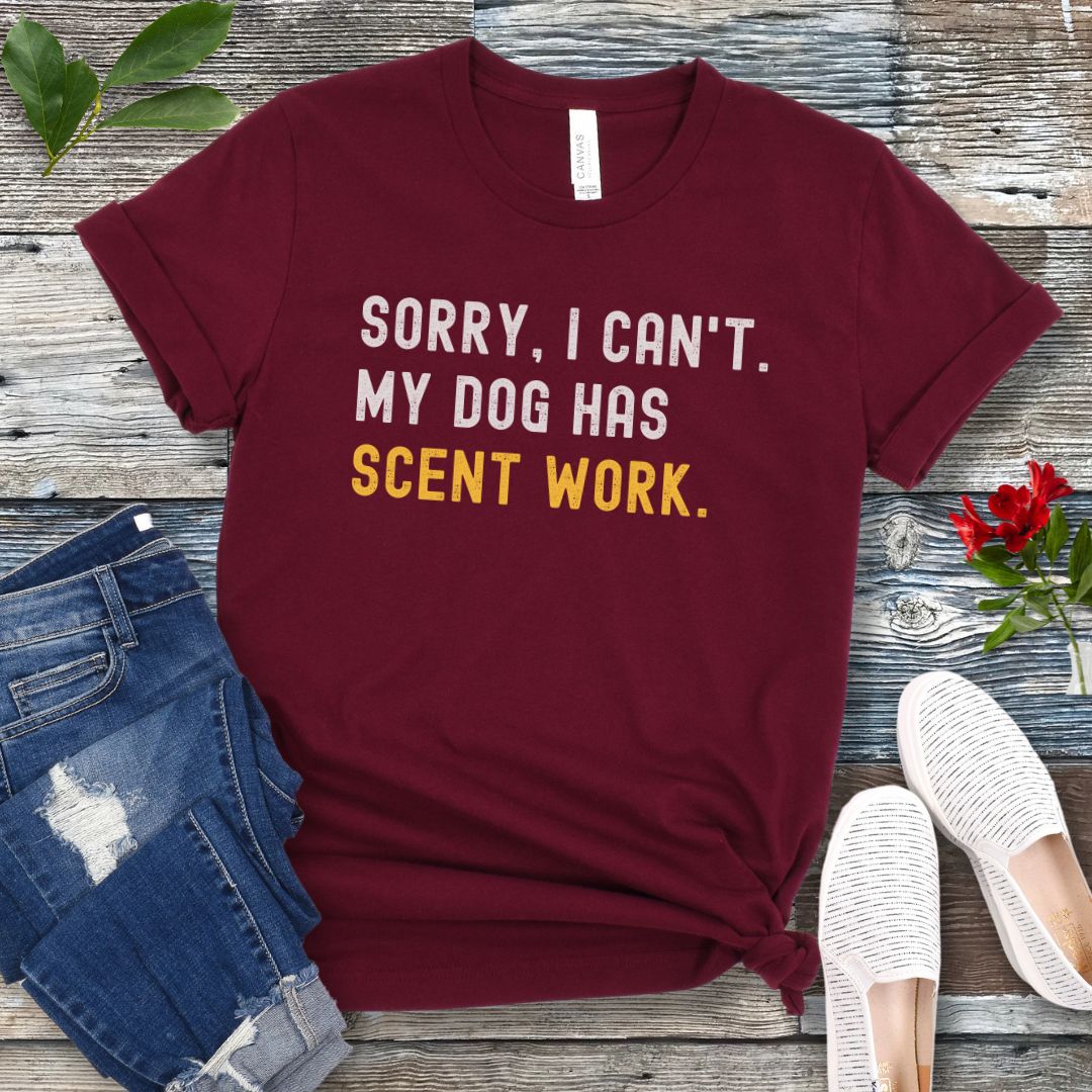 Maroon t-shirt with 'SORRY, CAN'T. MY DOG HAS SCENT WORK' text on a wooden surface with jeans and shoes.