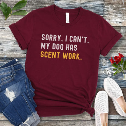 Maroon t-shirt with 'SORRY, CAN'T. MY DOG HAS SCENT WORK' text on a wooden surface with jeans and shoes.