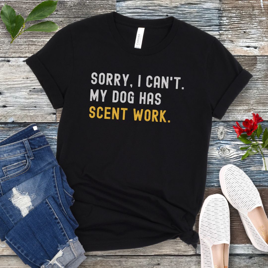 Black t-shirt with 'SORRY, CAN'T. MY DOG HAS SCENT WORK' text on a wooden surface with jeans and shoes.