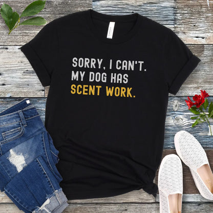 Black t-shirt with 'SORRY, CAN'T. MY DOG HAS SCENT WORK' text on a wooden surface with jeans and shoes.