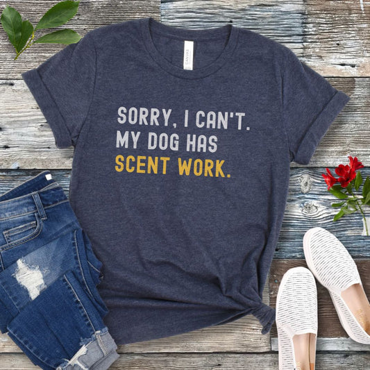 Heather navy blue t-shirt with 'SORRY, CAN'T. MY DOG HAS SCENT WORK' text on a wooden surface with jeans and shoes.