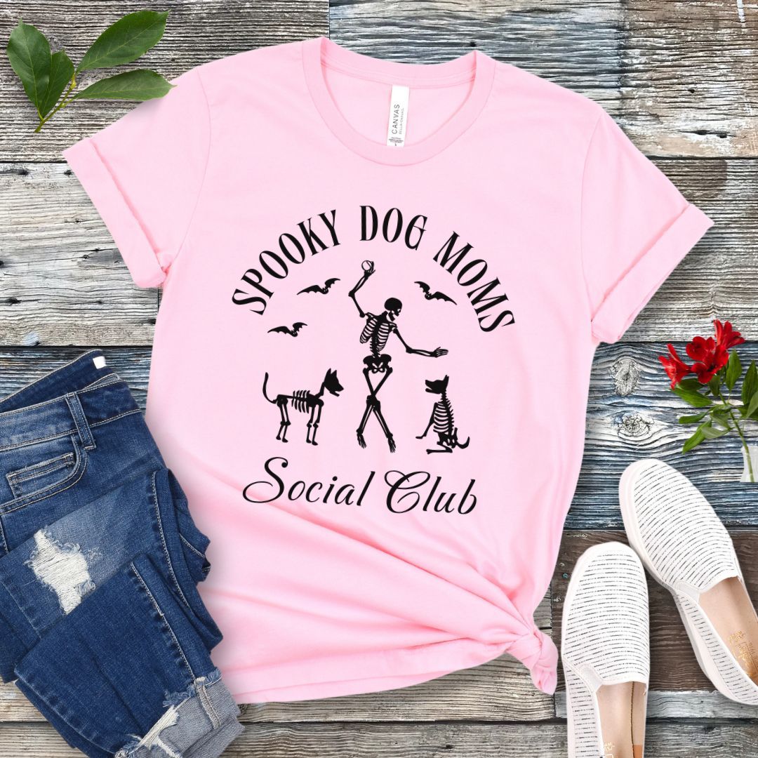 Pink t-shirt with 'Spooky Dog Moms Social Club' text and skeleton playing with skeleton dogs, presented on a wooden surface with jeans and shoes.