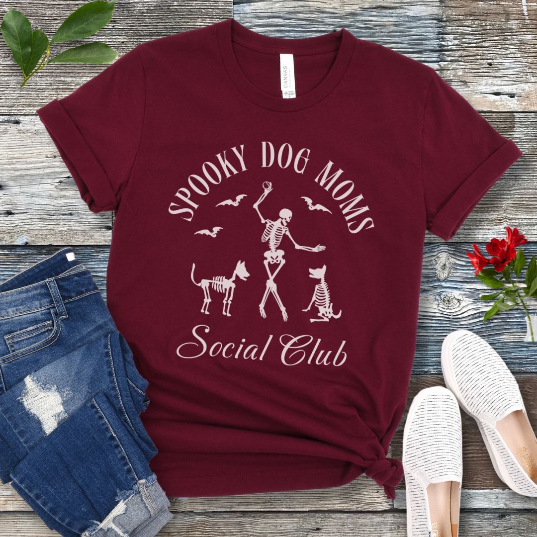 Maroon t-shirt with 'Spooky Dog Moms Social Club' text and skeleton playing with skeleton dogs, presented on a wooden surface with jeans and shoes.