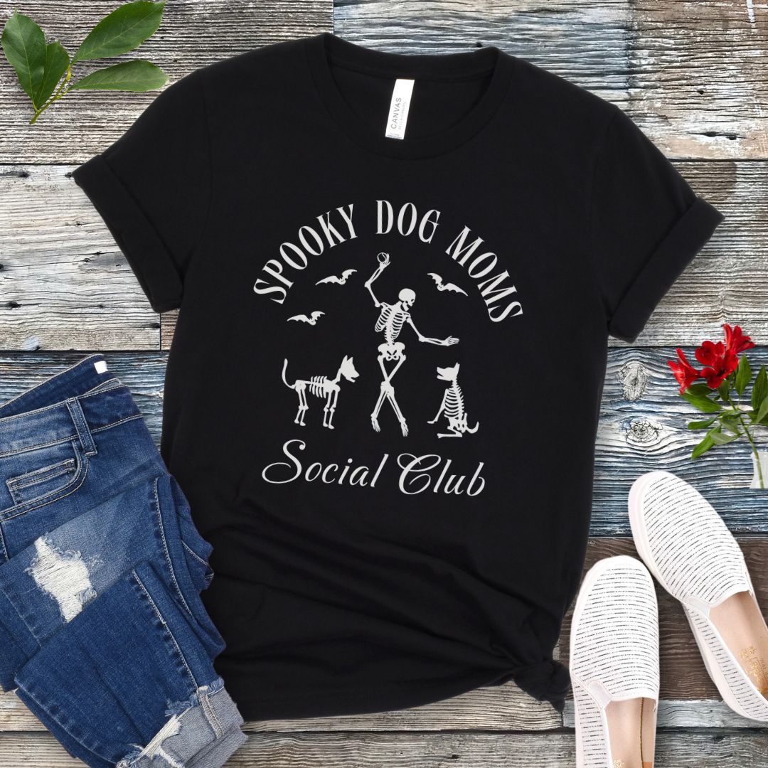 Black t-shirt with 'Spooky Dog Moms Social Club' text and skeleton playing with skeleton dogs, presented on a wooden surface with jeans and shoes.