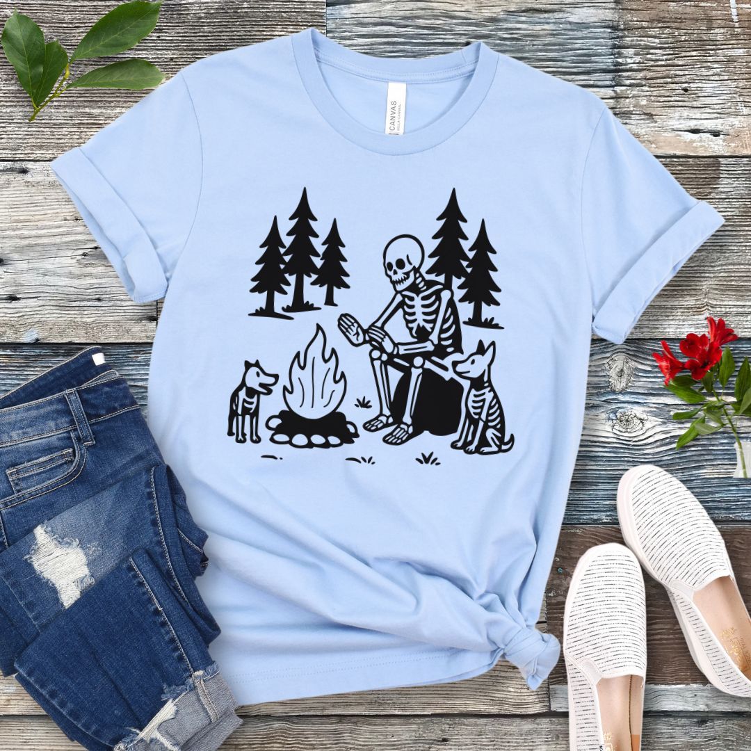 A light blue t-shirt for dog lovers with a graphic print of a skeleton human and two dogs roasting marshmallows over a campfire, surrounded by pine trees.