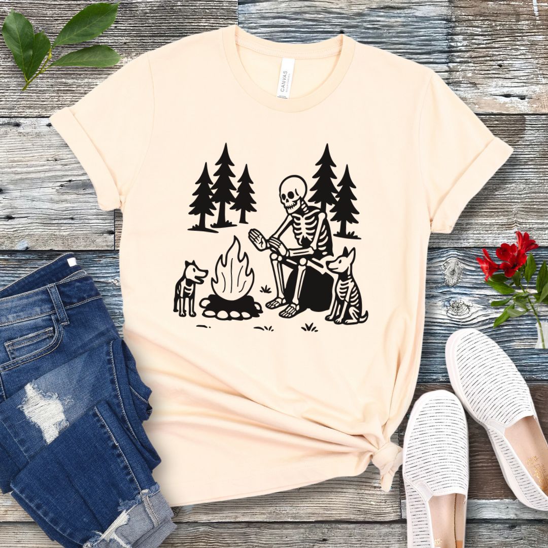 A beige t-shirt for dog lovers with a graphic print of a skeleton human and two dogs roasting marshmallows over a campfire, surrounded by pine trees.