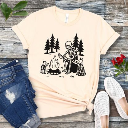 A beige t-shirt for dog lovers with a graphic print of a skeleton human and two dogs roasting marshmallows over a campfire, surrounded by pine trees.