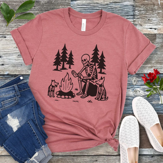 A mauve t-shirt for dog lovers with a graphic print of a skeleton human and two dogs roasting marshmallows over a campfire, surrounded by pine trees.