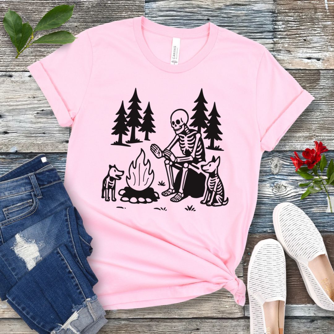 A pink t-shirt for dog lovers with a graphic print of a skeleton human and two dogs roasting marshmallows over a campfire, surrounded by pine trees.