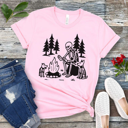 A pink t-shirt for dog lovers with a graphic print of a skeleton human and two dogs roasting marshmallows over a campfire, surrounded by pine trees.