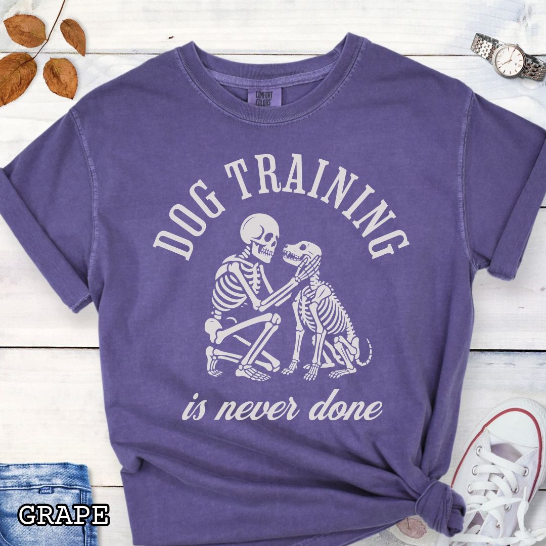 Purple Comfort Colors t-shirt for spooky dog lovers with skeleton dog and human and the text 'Dog training is never done' in vintage lettering, shown on a wooden surface.