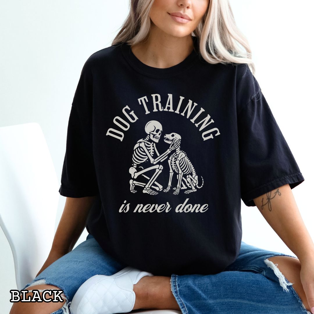 Black Comfort Colors t-shirt for spooky dog lovers with skeleton dog and human and the text 'Dog training is never done' in vintage lettering, shown on a wooden surface.