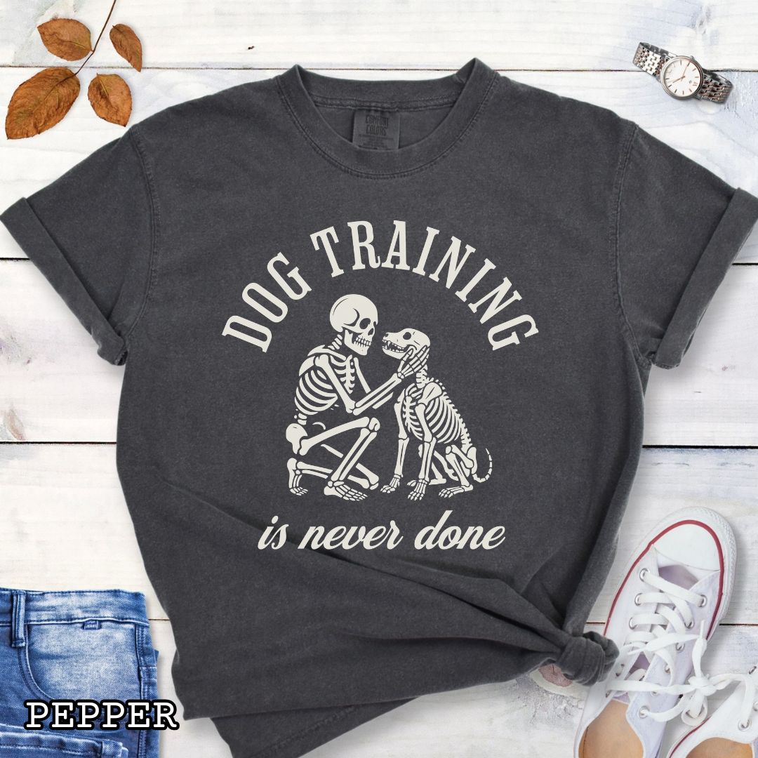 Dark grey Comfort Colors t-shirt for spooky dog lovers with skeleton dog and human and the text 'Dog training is never done' in vintage lettering, shown on a wooden surface.