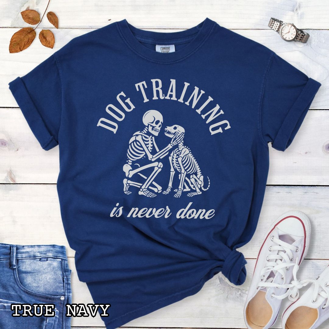 Navy Comfort Colors t-shirt for spooky dog lovers with skeleton dog and human and the text 'Dog training is never done' in vintage lettering, shown on a wooden surface.