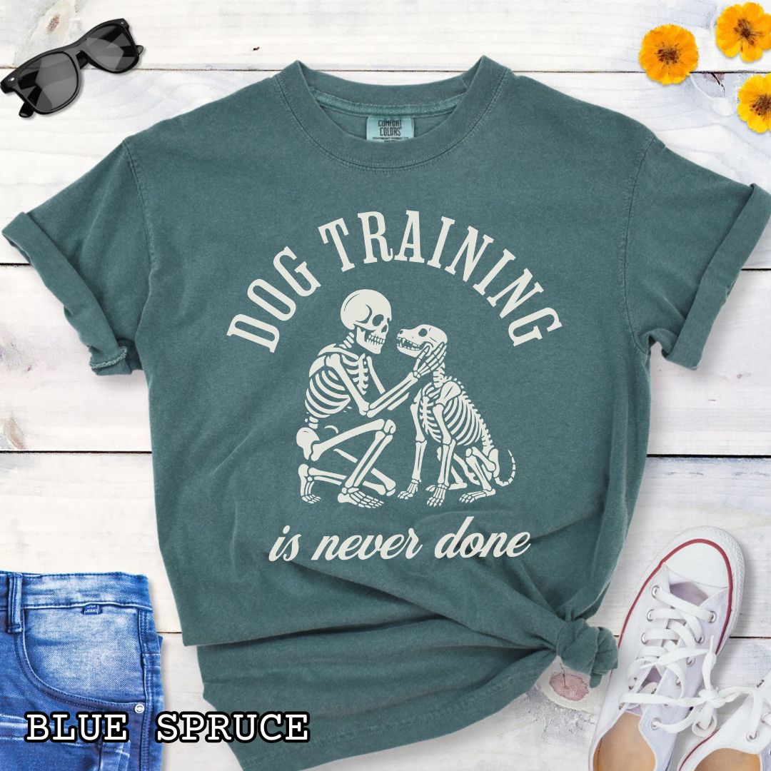 Green Comfort Colors t-shirt for spooky dog lovers with skeleton dog and human and the text 'Dog training is never done' in vintage lettering, shown on a wooden surface.