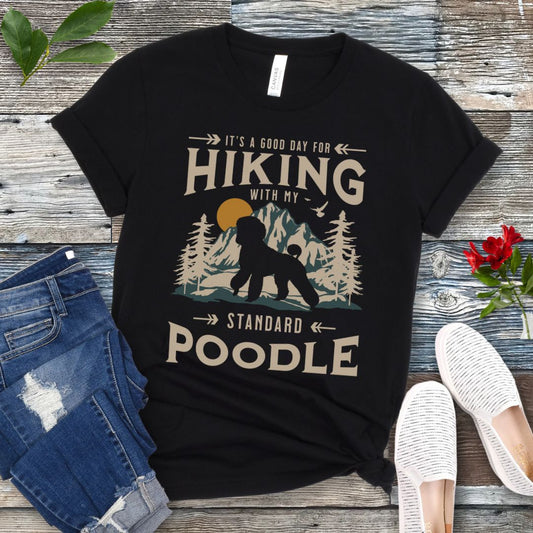 Black dog hiking t-shirt with 'Hiking with my Standard Poodle' text, mountain scene and Standard Poodle silhouette, presented on a wooden surface with jeans and shoes.