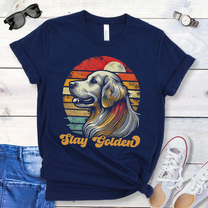 Navy t-shirt with 'Stay Golden' text and a vintage sunset graphic with a line drawn Golden Retriever, presented on a wooden surface with jeans and shoes.