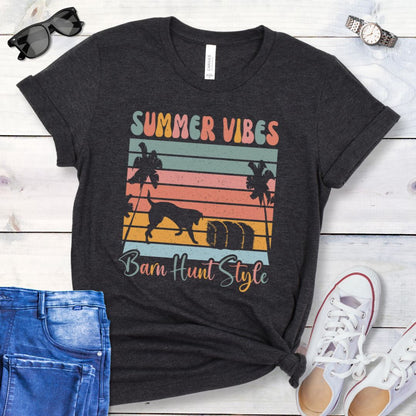 A dark grey barn hunt t-shirt with a graphic print of palm trees, vintage sunset stripes, a dog investigating a hale bale, and multicolor text reading 'SUMMER VIBES Barn Hunt Style', displayed on a wooden surface with a pair of blue jeans and white shoes in the background.
