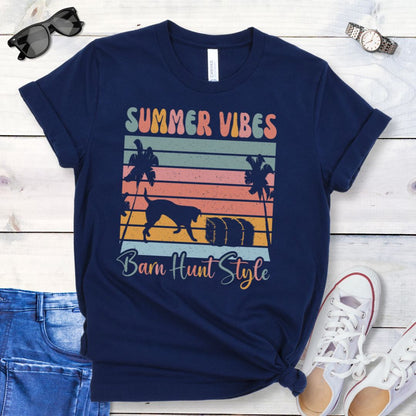 A navy blue barn hunt t-shirt with a graphic print of palm trees, vintage sunset stripes, a dog investigating a hale bale, and multicolor text reading 'SUMMER VIBES Barn Hunt Style', displayed on a wooden surface with a pair of blue jeans and white shoes in the background.