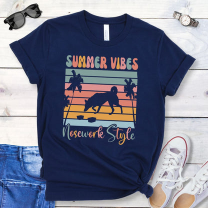 Navy t-shirt with vintage sunset, sniffing dog and text 'Summer Vibes Nosework Style' design on a wooden surface with jeans and shoes.