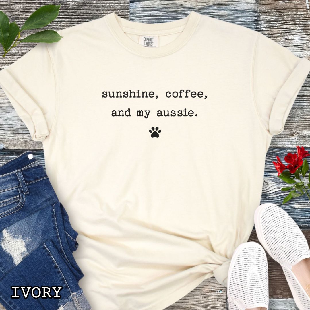 Beige Comfort Colors t-shirt for Australian Shepherd lovers with 'sunshine, coffee, and my aussie' text in minimalist typewriter lettering and paw print, shown on a wooden surface with accessories.