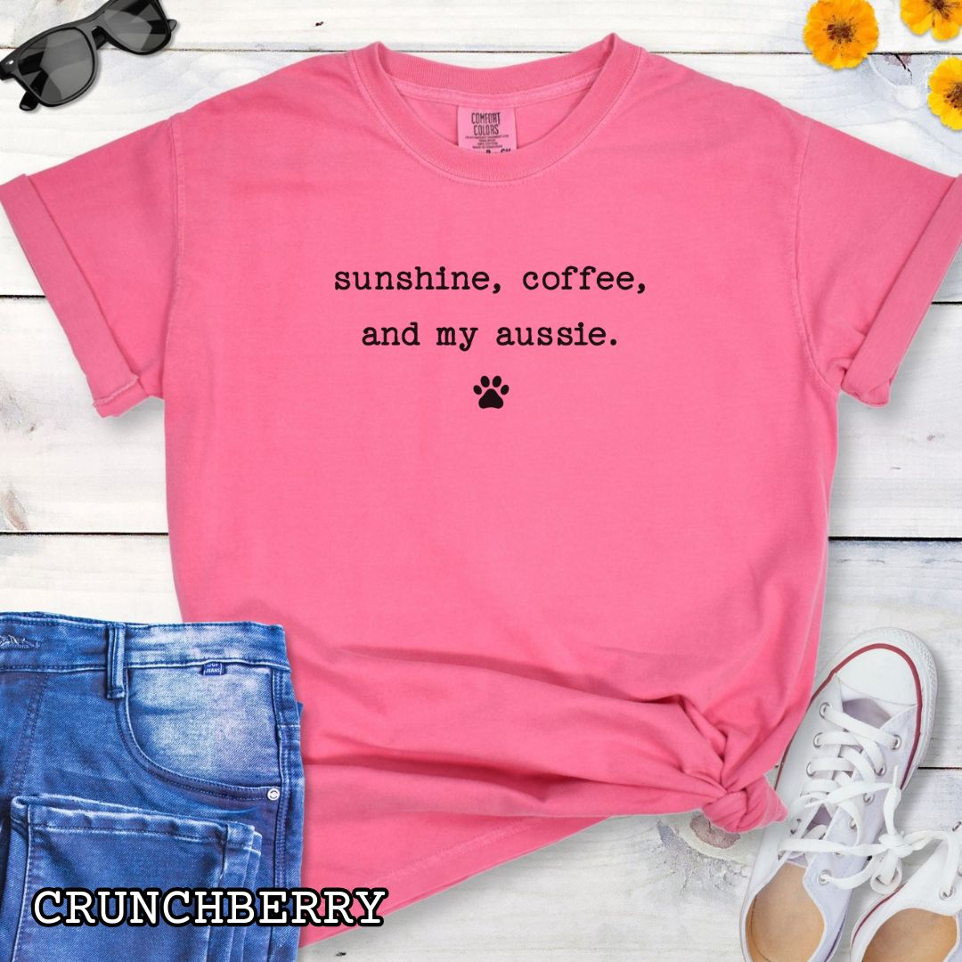 Pink Comfort Colors t-shirt for Australian Shepherd lovers with 'sunshine, coffee, and my aussie' text in minimalist typewriter lettering and paw print, shown on a wooden surface with accessories.