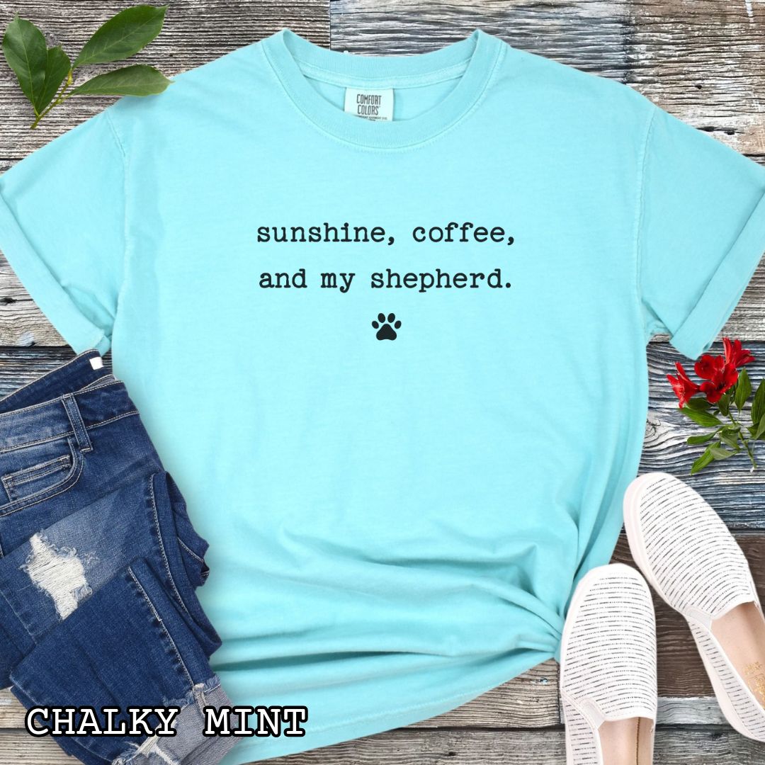 Mint Comfort Colors t-shirt for shepherd moms with 'sunshine, coffee, and my shepherd' text in minimalist typewriter lettering and paw print, shown on a wooden surface with accessories.