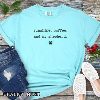 Mint Comfort Colors t-shirt for shepherd moms with 'sunshine, coffee, and my shepherd' text in minimalist typewriter lettering and paw print, shown on a wooden surface with accessories.
