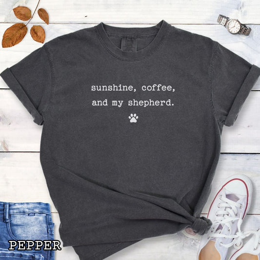 Dark grey Comfort Colors t-shirt for German Shepherd lovers with 'sunshine, coffee, and my shepherd' text in minimalist typewriter lettering and paw print, shown on a wooden surface with accessories.