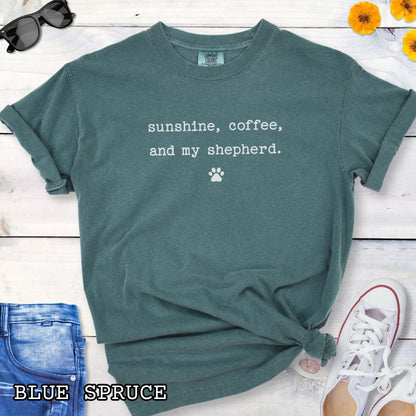 Green Comfort Colors t-shirt for German Shepherd lovers with 'sunshine, coffee, and my shepherd' text in minimalist typewriter lettering and paw print, shown on a wooden surface with accessories.