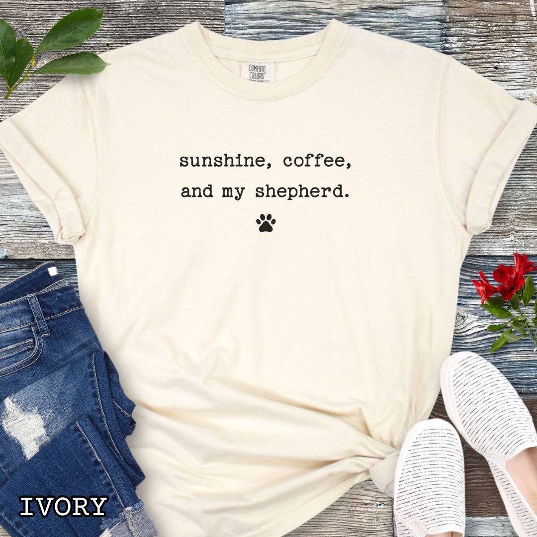 Beige Comfort Colors t-shirt for German Shepherd owners with 'sunshine, coffee, and my shepherd' text in minimalist typewriter lettering and paw print, shown on a wooden surface with accessories.